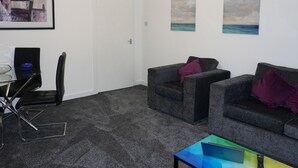 A modern living room with two grey sofas, a glass coffee table, and a black chair. There are two framed paintings on the wall, a white door, and a ceiling light.