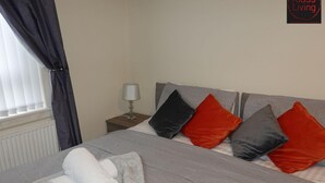A neatly made bed with grey bedding and multiple pillows in black and red. There is a lamp on a bedside table and a towel on the bed.