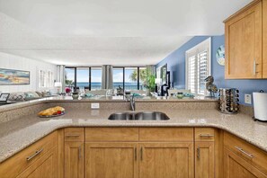 A kitchen with a large window, a sink, wooden cabinets, and a dining area with a view of the ocean.