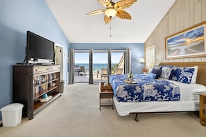 A bedroom with a bed, a television, a bookshelf, a ceiling fan, and a view of the outdoors.