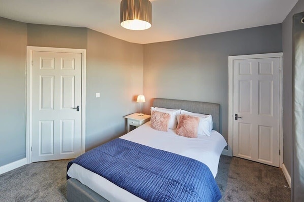 A bedroom with a bed, two white doors, a nightstand with a lamp, and a ceiling light fixture.