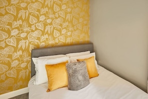 Una cama con ropa blanca, un plaid suave gris y dos almohadas amarillas.