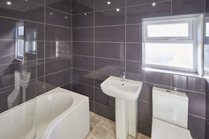 Un baño moderno con paredes de azulejos oscuros, una bañera blanca, lavamanos pedestal y ventana que deja entrar luz natural.