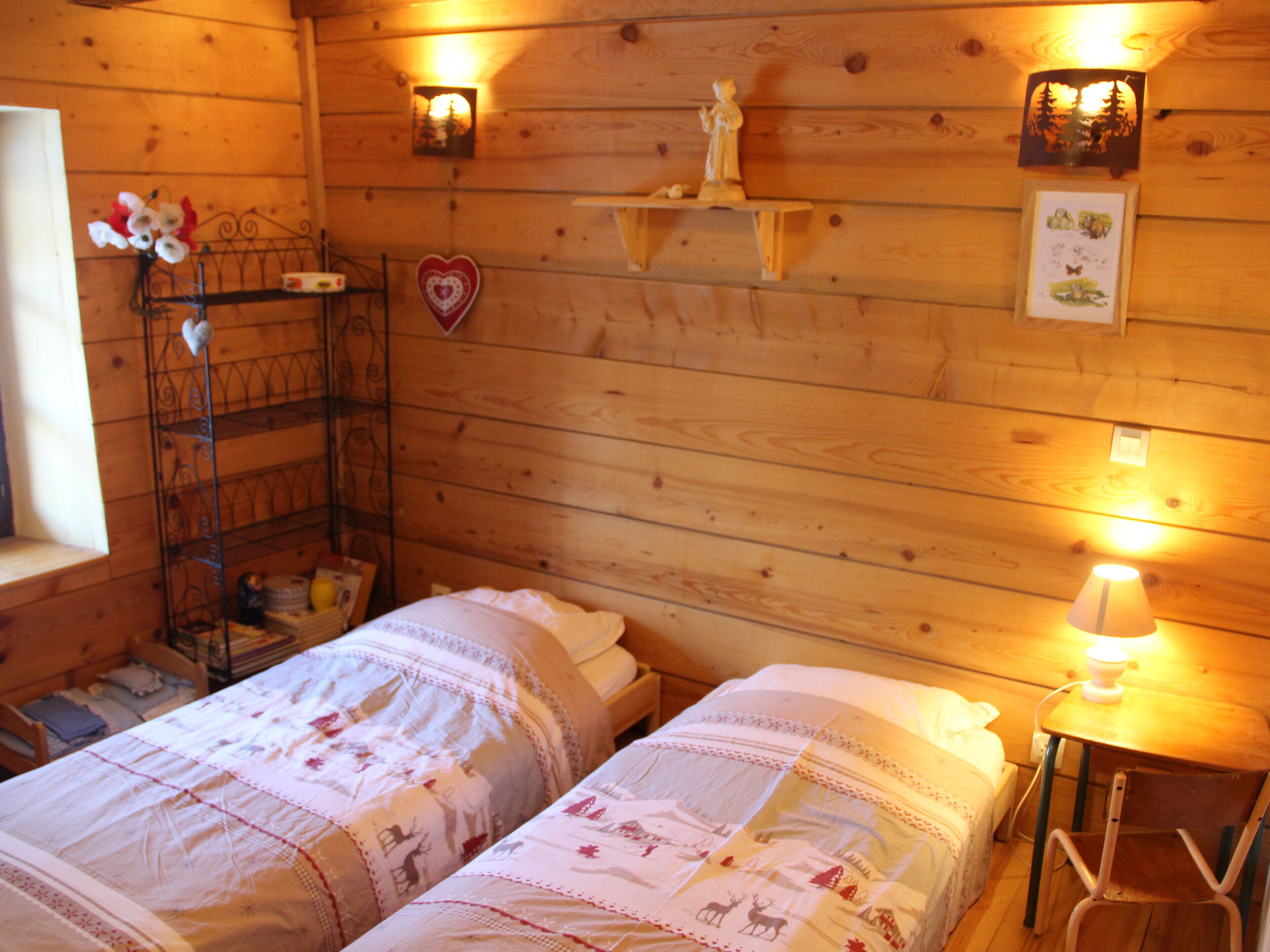 Una habitación acogedora con dos camas, una mesita con lámpara, una estantería con objetos decorativos y una ventana con cortinas.