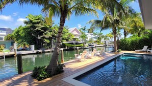 Zona de piscina con sillones de descanso, una terraza de madera y una casa al fondo.