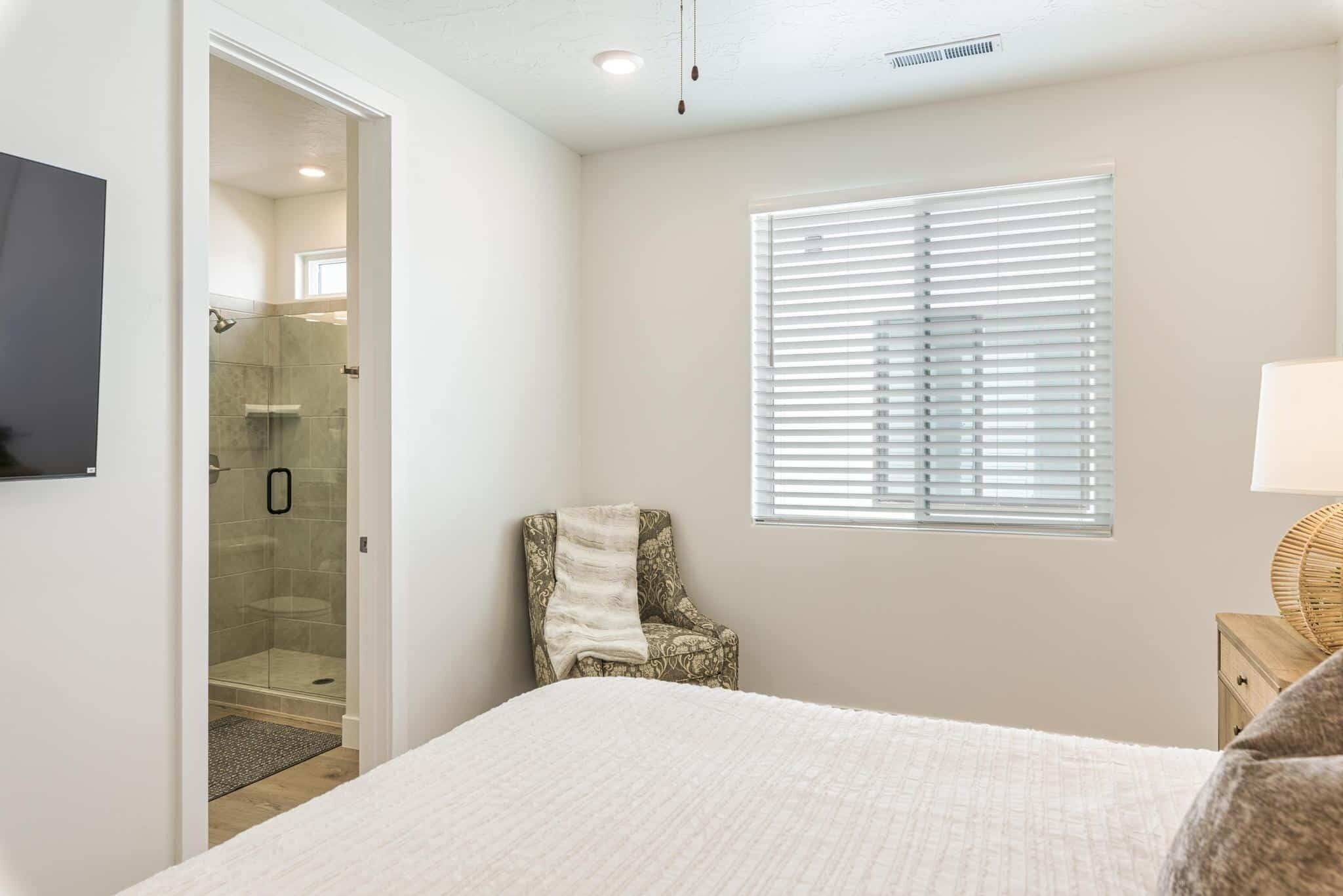 A bedroom with a bed, a chair, a window with blinds, a bedside table, and a TV mounted on the wall.