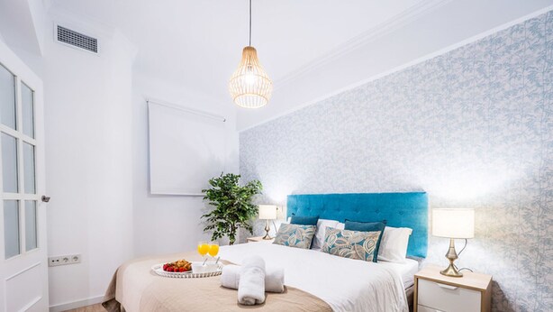 A hotel room with a large bed, a bedside table, a nightstand, a hanging light fixture, a wall-mounted air vent, and a window with blinds.