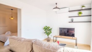 Una sala de estar moderna con un sofá color beige, un televisor de pantalla plana, una mesa de centro de madera y una planta en maceta. Se observan estantes con adornos y una lámpara colgante sobre el sofá.