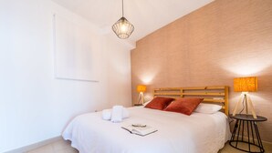 Una cama bien hecha con sábanas blancas y dos almohadas, una mesita de noche con lámpara y un aplique de pared.