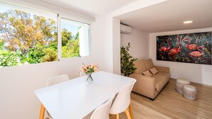 Ett modernt vardagsrum med ett vitt matbord, en beige soffa och ett stort fönster med utsikt över träd.