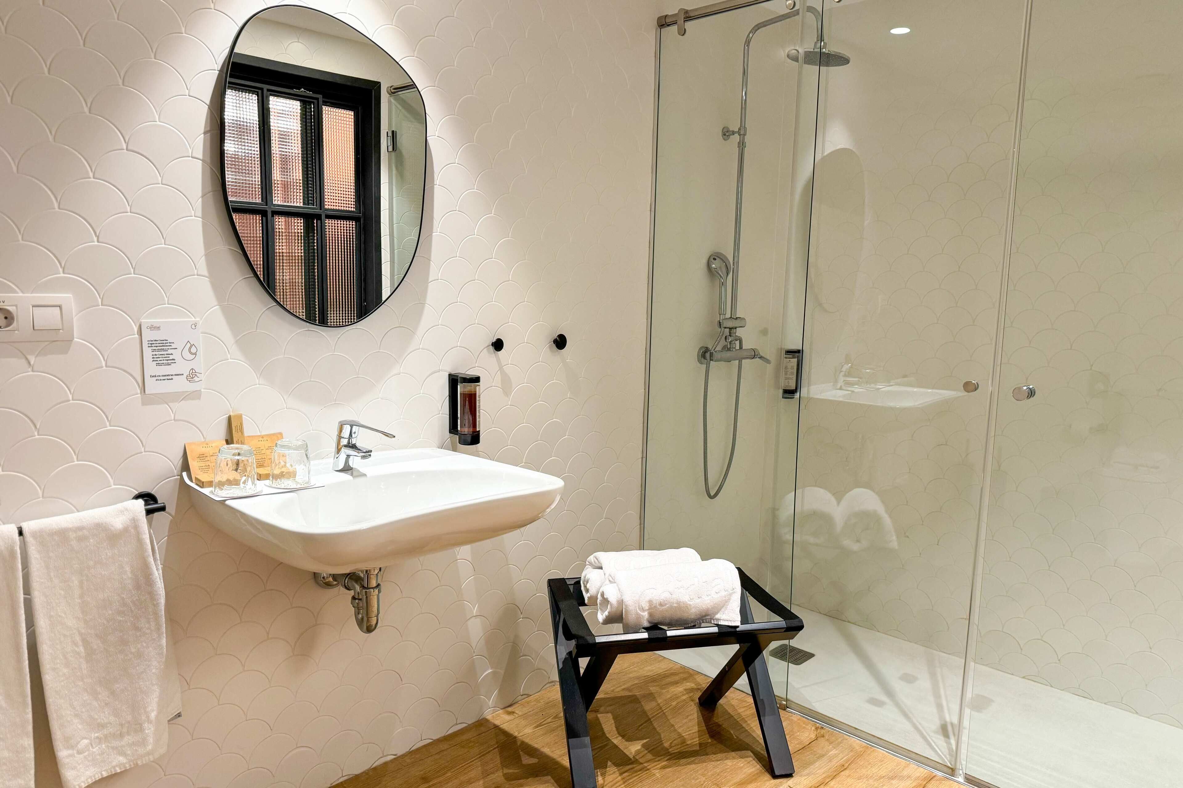 A bathroom with a white sink, a mirror, a glass shower enclosure, and a small black stool with a towel.