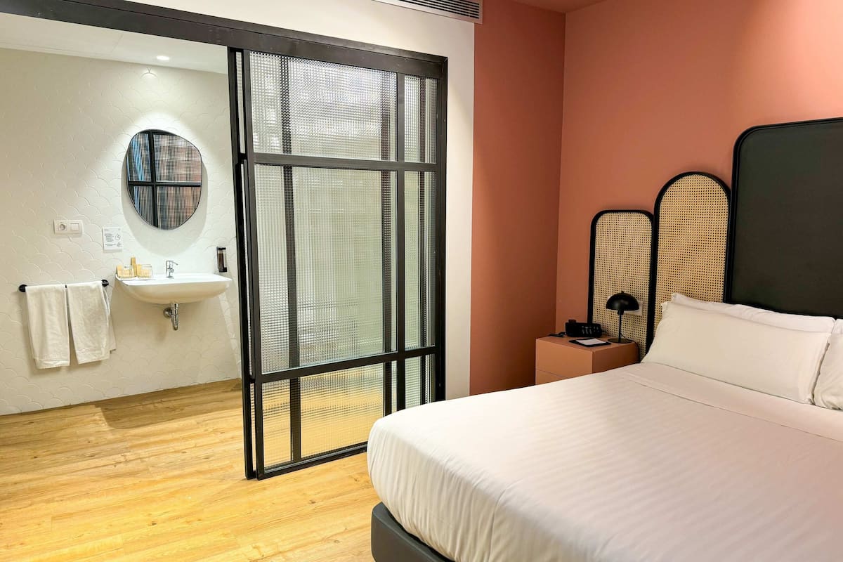 A hotel room with a bed, a bedside table with a lamp, a window with a view, a round mirror, and a sink.
