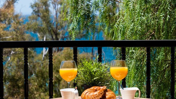 A table with croissants, two glasses of orange juice, and white cups on a wooden table outdoors.