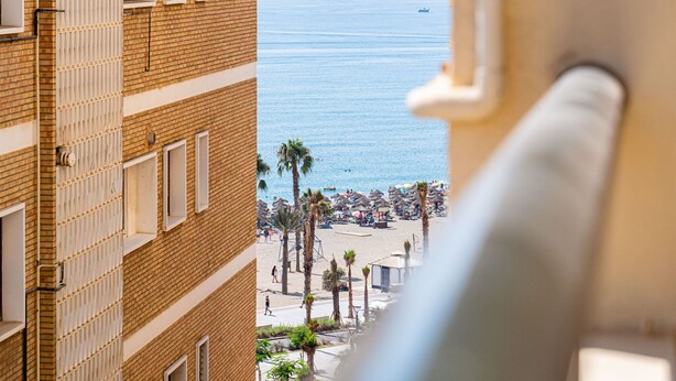 A view of a beach with palm trees and umbrellas, a boat in the distance, and a building with a brick facade and windows. - Sunset Malaga Apartments - Maestranza 8d2 (Málaga)