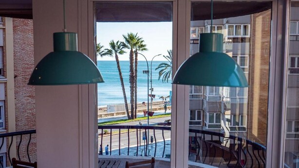 A balcony with a view of the beach and palm trees, featuring two green pendant lights and wooden furniture.