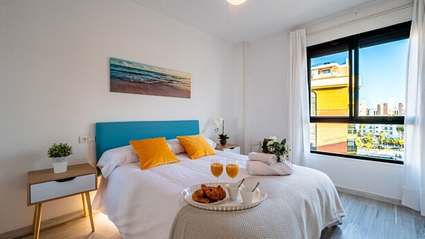 A modern hotel room with a bed, a bedside table with a plant, a window with a city view, and a painting on the wall. - Sunset Malaga Apartments - PALM BEACH (Torremolinos)