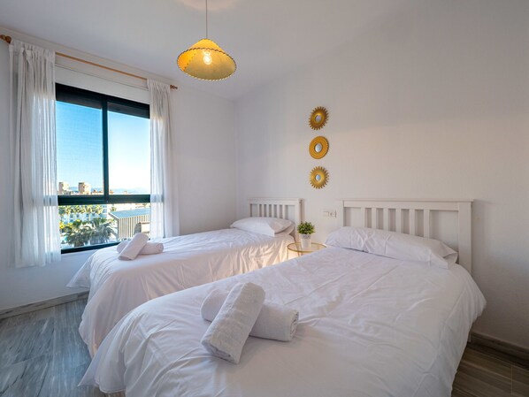Habitación de hotel con dos camas, un ventanal con cortinas, una lámpara colgante y espejos decorativos en la pared.