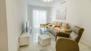 Ein modernes Wohnzimmer mit einem Glastisch, einem beigefarbenen Sofa mit orangefarbenen Kissen, einem braunen Ledersessel und einem Flachbildfernseher auf einem weißen Lowboard.