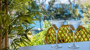 Two martini glasses with olives on a table with a yellow lattice chair, set against a backdrop of greenery and a body of water.