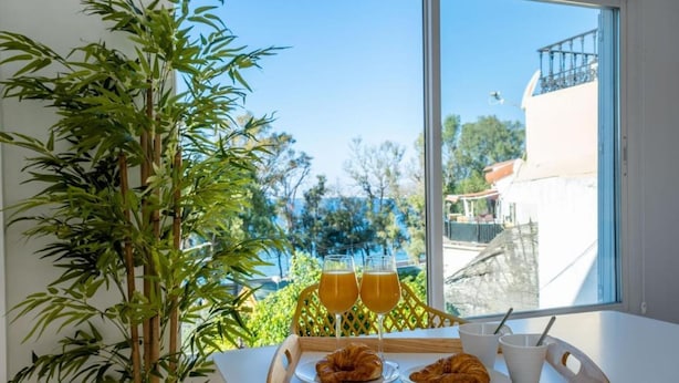 A breakfast tray with croissants and two glasses of orange juice on a table by a window overlooking a balcony with a view of trees and buildings.