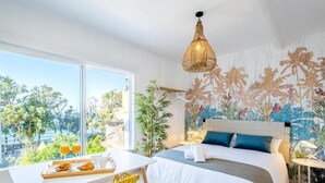 A hotel room with a bed, a dining table with breakfast items, a large window with a view of palm trees, and a tropical-themed wall mural.