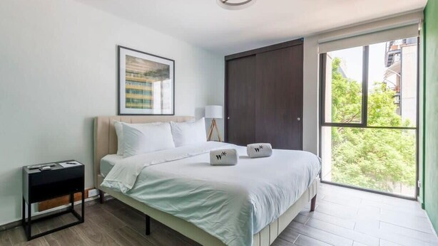 Een moderne hotelkamer met een bed, nachtkastje, lamp, een ingelijste foto aan de muur en een groot raam met uitzicht op groen.