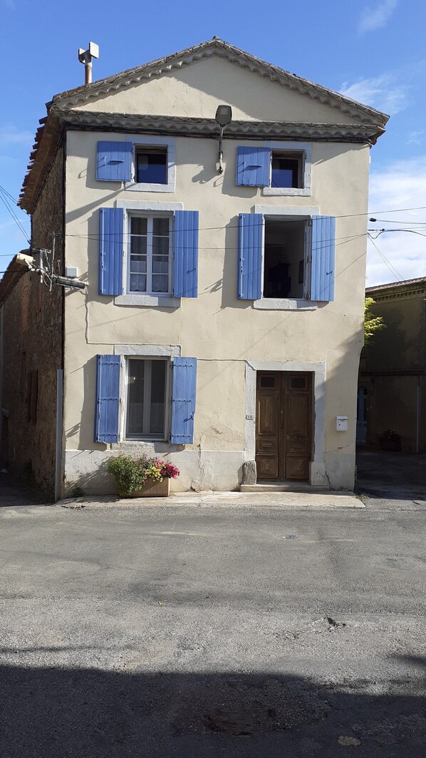 Un bâtiment à deux étages avec des volets bleus, une porte en bois et un petit jardinier devant l’entrée.