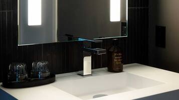 Une salle de bain moderne avec un grand miroir, un lavabo blanc, une bouteille de shampoing et deux verres.