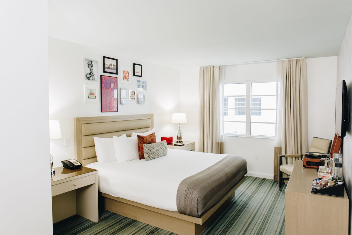 A hotel room with a large bed, a desk with a lamp, a chair, a TV, and a window with curtains.