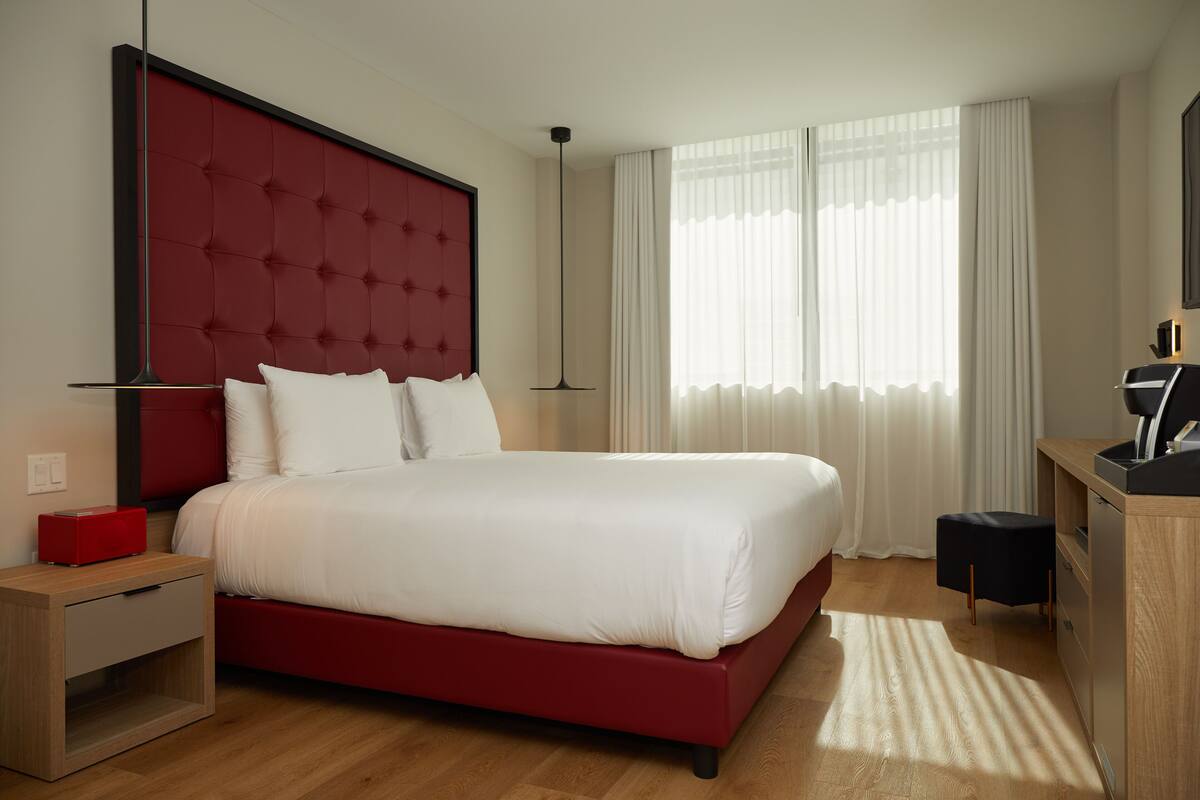 A hotel room with a red upholstered bed, a wooden nightstand, a desk with a chair, and a large window with curtains.