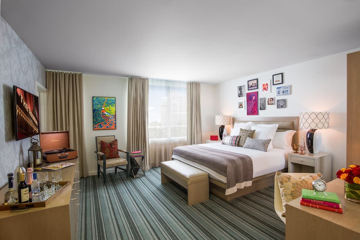 A hotel room with a bed, bedside tables, a desk with a lamp, a chair, a bench, a TV, and a window with curtains.