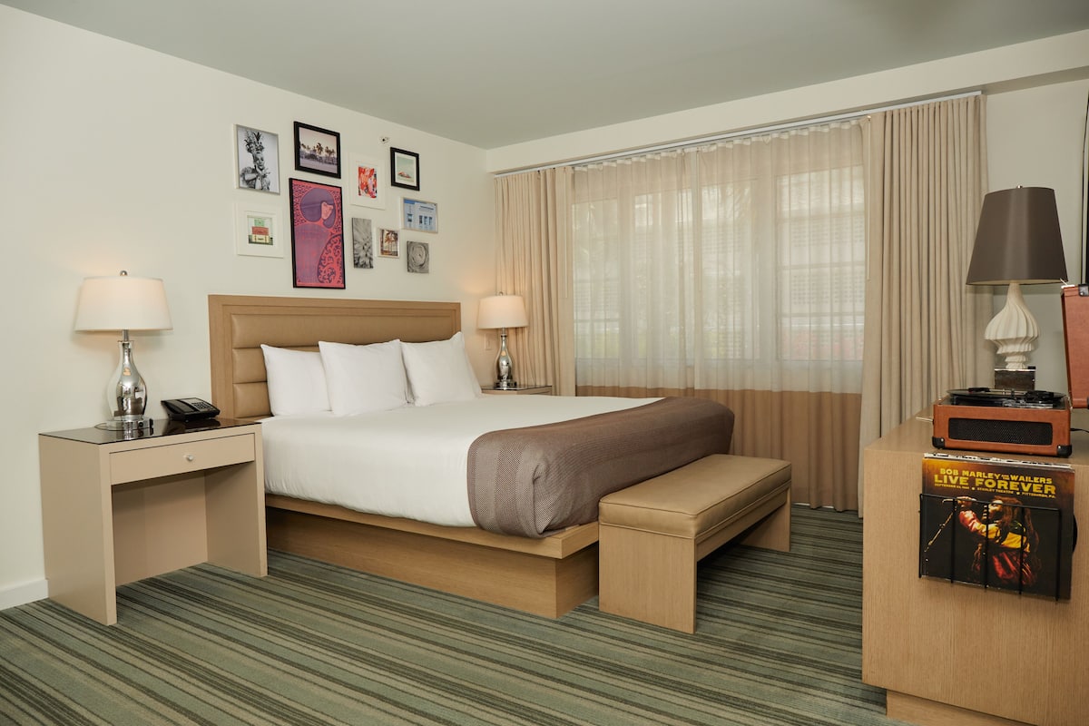 A hotel room with a bed, bedside tables, a bench, a lamp, a poster, and a window with curtains.