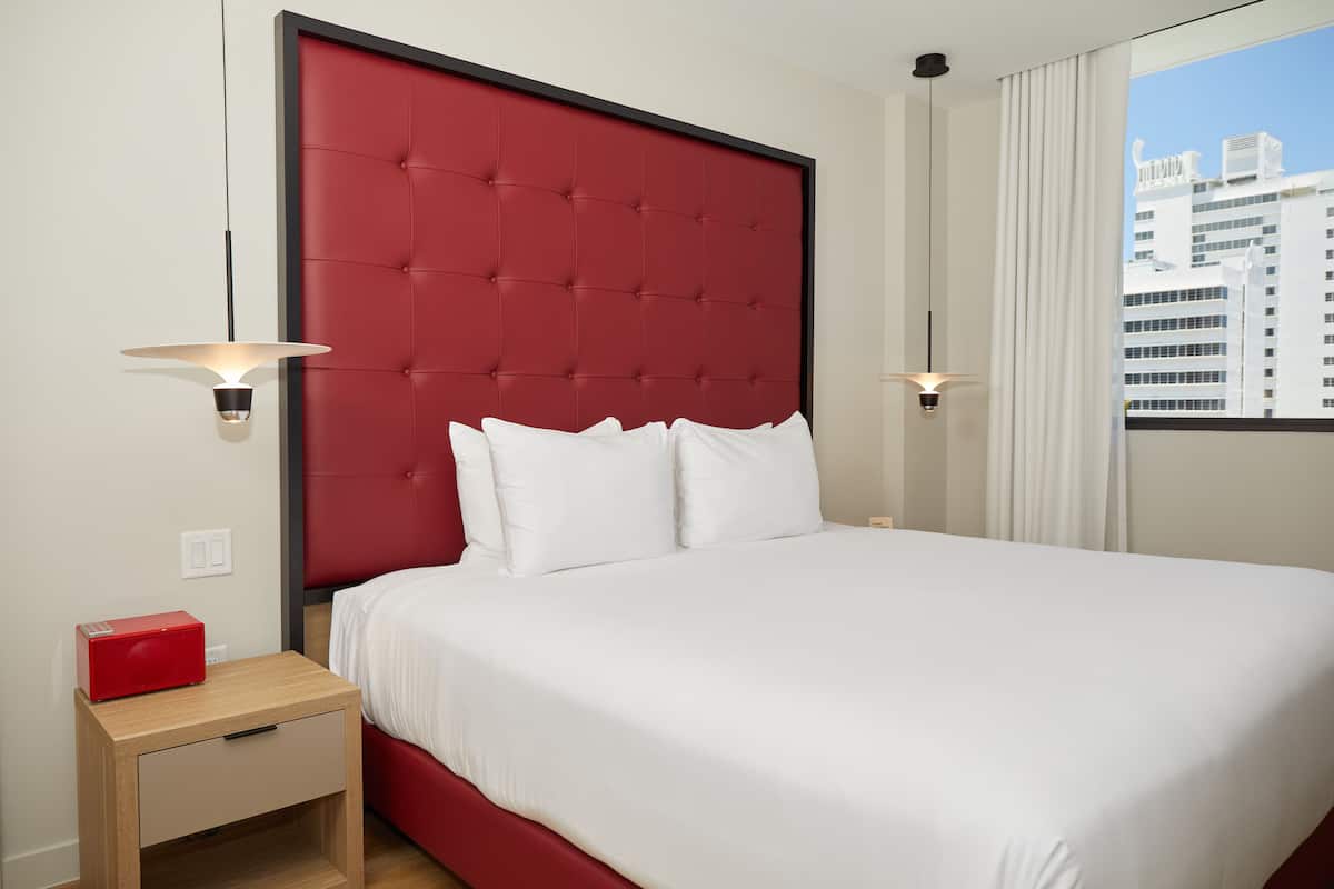 A hotel room with a red headboard, white bedding, two bedside lamps, a nightstand with a red radio, and a window with a city view.