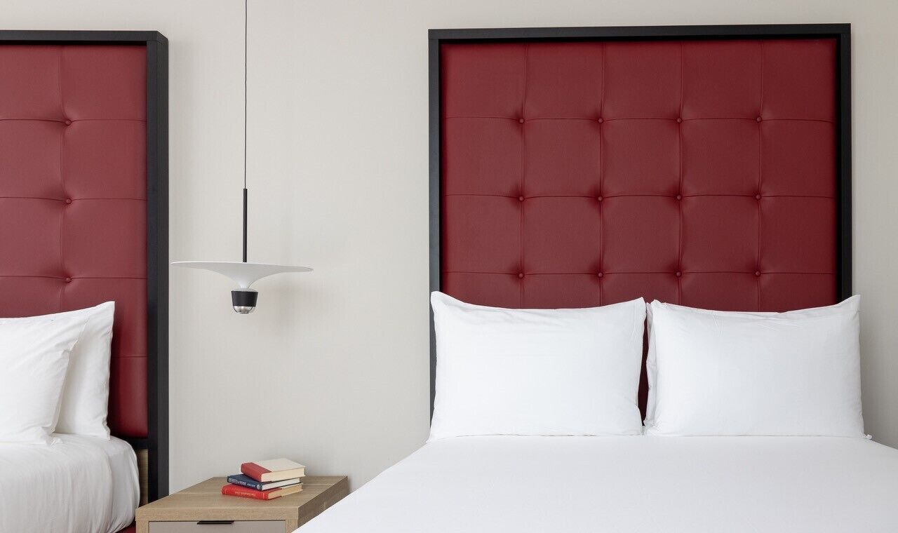 A hotel room with two beds, each with a red headboard, white bedding, and a bedside table with books.