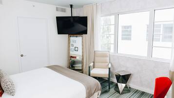 A hotel room with a bed, a TV mounted on the wall, a chair, a small table, and a window with a view of buildings.
