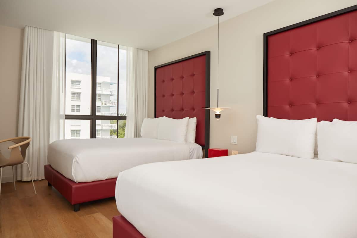 A hotel room with two beds, a red headboard, a window with curtains, a small table, and a chair.