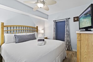 Un lit bien fait, avec une tête de lit en bois, un ventilateur de plafond, un téléviseur posé sur une commode en bois, et une vue sur l’océan par la fenêtre.