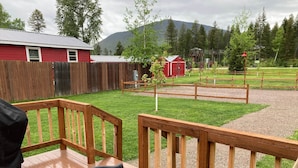 A wooden deck with a table and chairs, a red building, and a fenced area with trees and a mountain in the background.