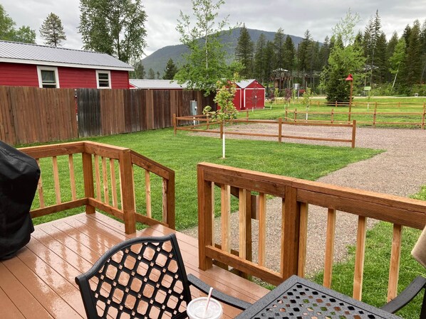 Une terrasse en bois avec une table et des chaises, un bâtiment rouge et une zone délimitée par une clôture, avec des arbres et une montagne en arrière-plan.