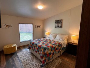 Une chambre à coucher avec un lit, deux lampes de chevet, une fenêtre avec des stores, un tapis et un petit pouf rond.