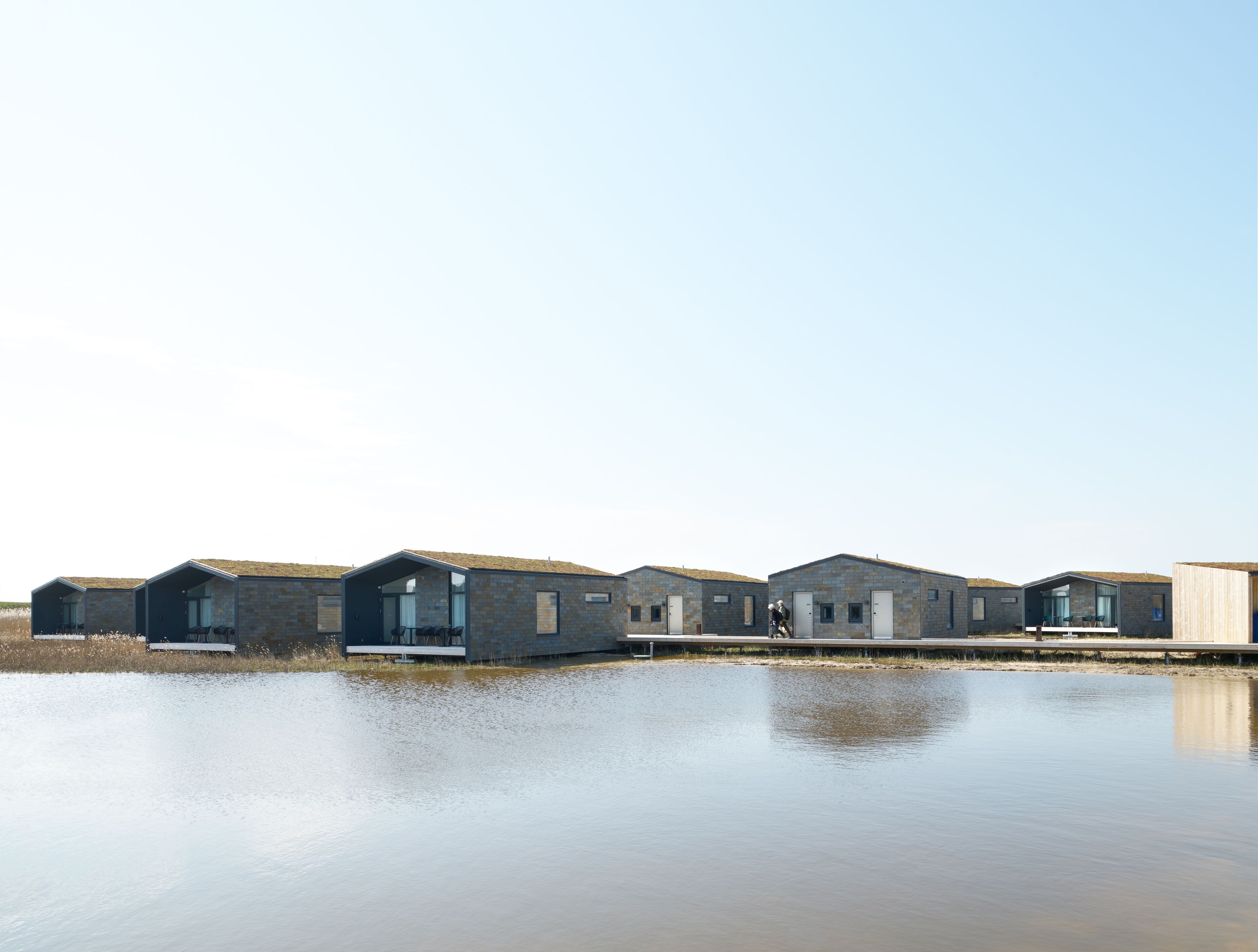 A row of modern, brick-roofed buildings with large windows, situated by a calm body of water.