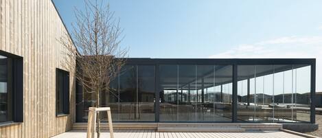 A modern building with large glass windows, a wooden deck, and a tree.