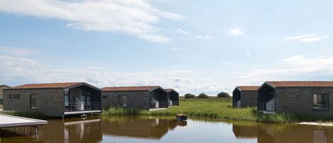 A row of modern, stone-roofed cabins with glass doors, situated by a calm body of water, reflecting the sky and surrounding vegetation.