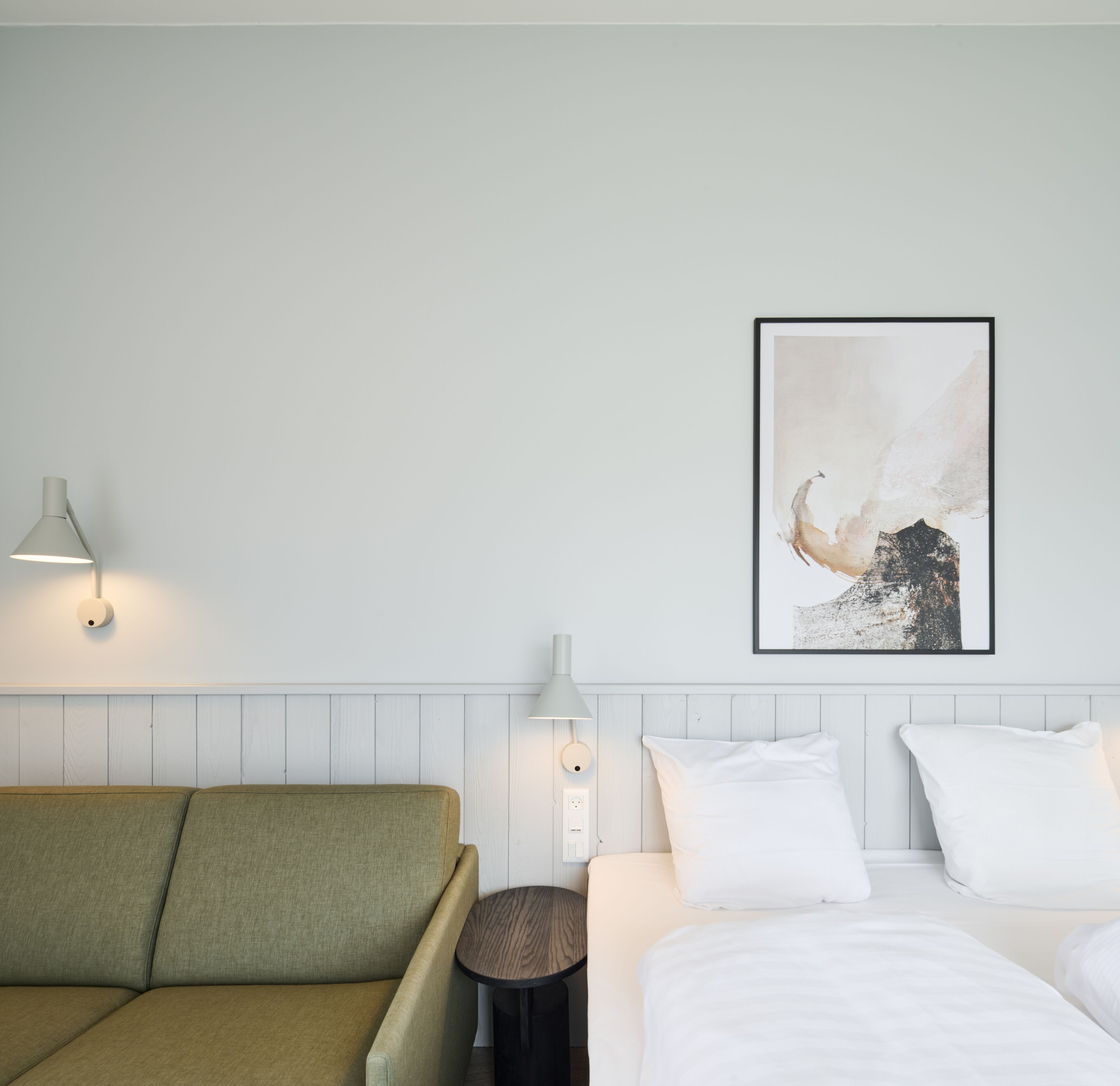 Habitación de hotel con sofá verde, cama con ropa de cama blanca, mesita de madera redonda y un cuadro abstracto colgado en la pared.