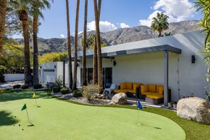 A modern building with a patio area, a golf putting green, and a mountain backdrop.