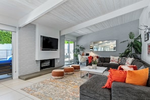 A modern living room with a flat-screen TV, grey sofa, orange cushions, and a patterned rug. - Far Far Away in Vista Las Palmas–Resort Pool Home (Palm Springs)