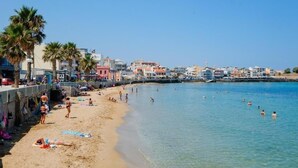 A beach with people swimming and sunbathing, palm trees, and buildings in the background. - Melina Apartments (Chania)