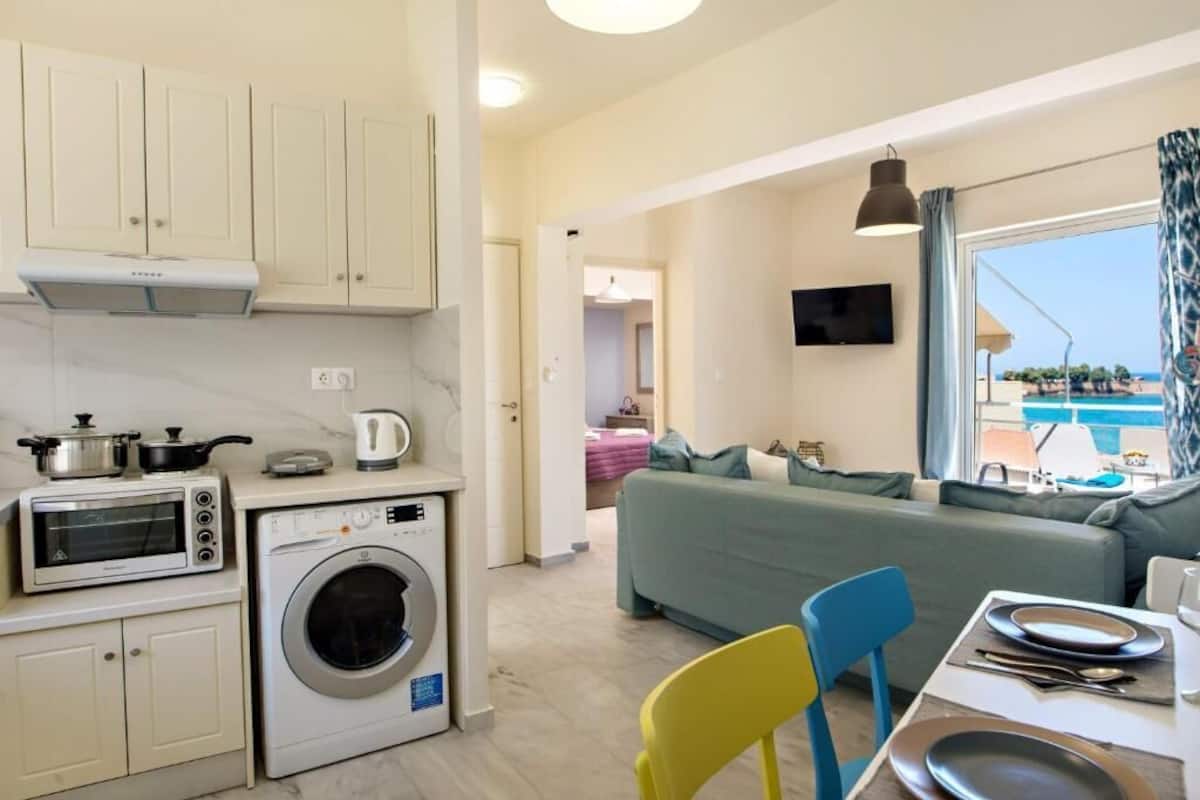 A compact living space with a kitchenette, a washing machine, a dining area with chairs, and a view of the sea.