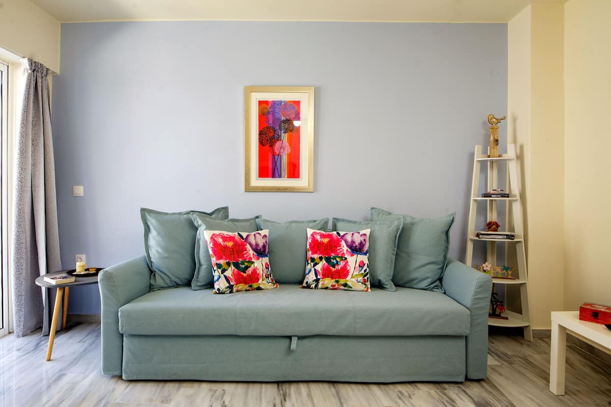 A living room with a light blue sofa, two floral throw pillows, a framed artwork on the wall, a white bookshelf, and a small table with a red box.