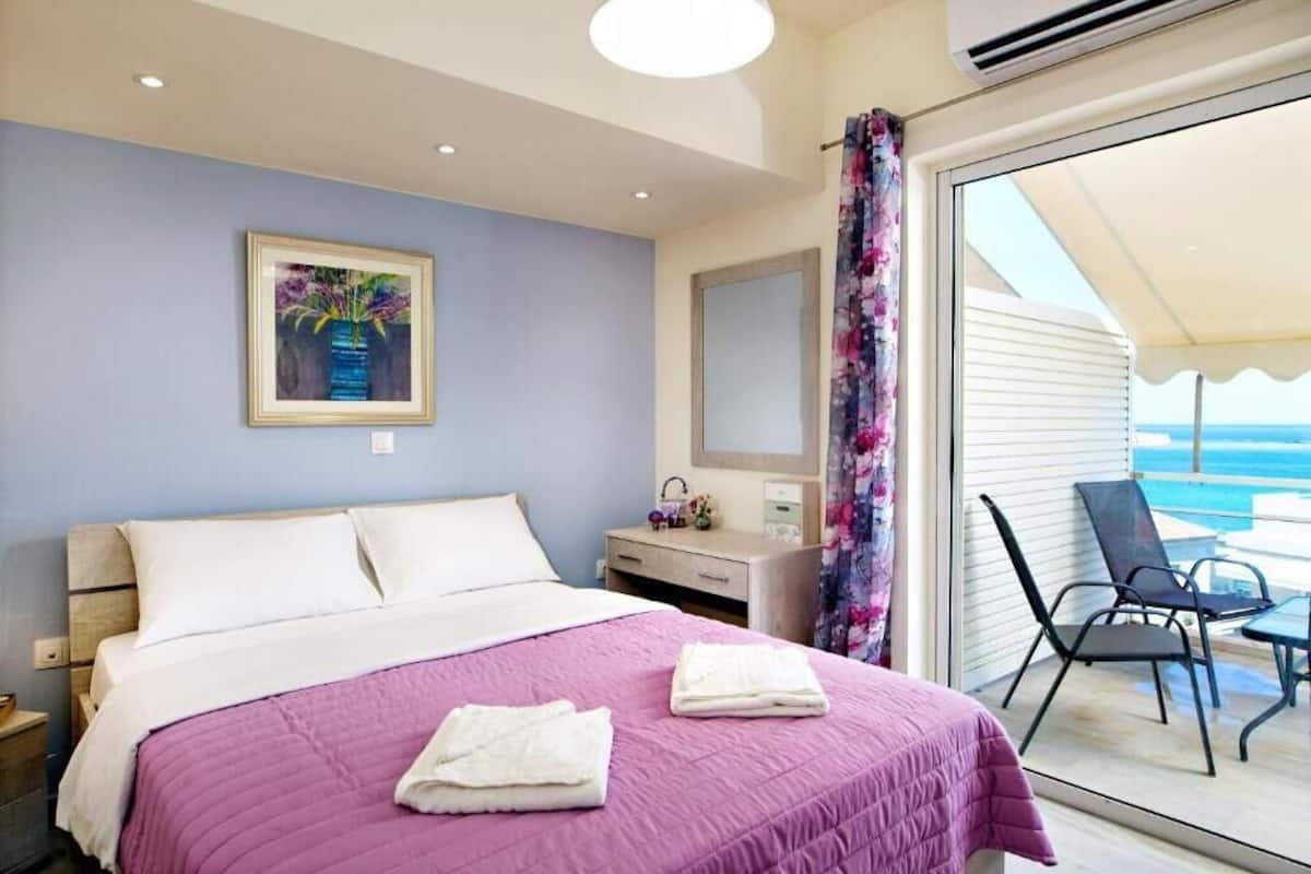 A bedroom with a bed, a dresser, a mirror, and a view of the balcony with chairs.